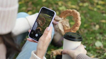 In autumn, two cheerful girls walk through the park during the day, drink coffee and take pictures of glasses of coffee and bagels against the background of autumn foliage for social networks.の写真素材