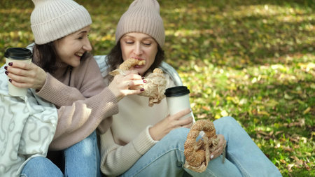 In autumn, two cheerful girls walk through the park during the day, drink coffee and take pictures of glasses of coffee and bagels against the background of autumn foliage for social networks.の写真素材