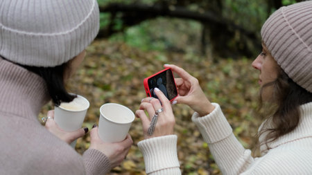 In autumn, two cheerful friends walk through the park during the day, drink coffee and take pictures of glasses of coffee against the background of autumn foliage for social networksの写真素材