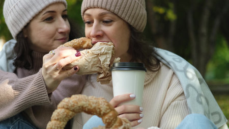 Two cute women in warm clothes sit hugging in an autumn park. Girlfriends enjoying beautiful weather, drinking tea or coffee, eating fresh bagels from the bakeryの写真素材