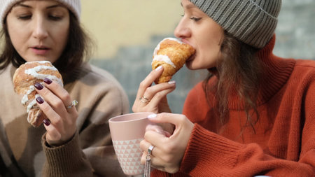 In autumn, two women drink tea from a thermos and eat croissants on the steps of the stairs. Friends in warm autumn clothes chatting outdoorsの写真素材