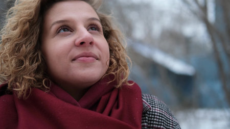 A young woman with curly hair raises her head and looks at the sky on a winter day. The woman is...
