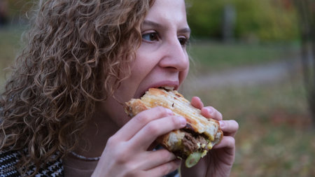 Young woman with curly hair bites and eats an appetizing delicious hot sandwich outdoors. Dressed break in autumn park. Likes to eat unhealthy fast food. A person enjoys food. Leisure in nature. Picnic.の写真素材