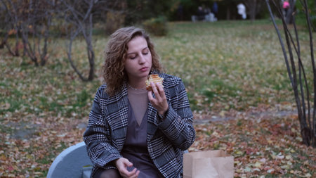 Young woman with curly hair bites and eats an appetizing delicious hot sandwich outdoors. Dressed break in autumn park. Likes to eat unhealthy fast food. A person enjoys food. Leisure in nature. Picnic.の写真素材