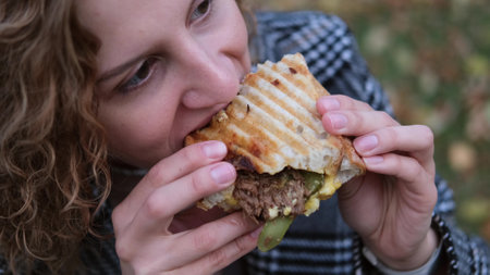 Young woman with curly hair bites and eats an appetizing delicious hot sandwich outdoors. Dressed break in autumn park. Likes to eat unhealthy fast food. A person enjoys food. Leisure in nature. Picnic.の写真素材