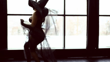Halloween theme: a young beautiful woman in a black gothic dress and a black witch hat dances near a large windowの写真素材