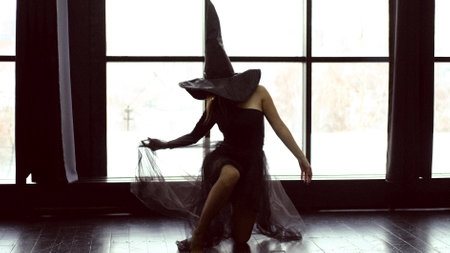 Halloween theme: a young beautiful woman in a black gothic dress and a black witch hat dances near a large windowの写真素材