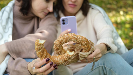 In autumn, two cheerful girls walk through the park during the day, drink coffee and take pictures of glasses of coffee and bagels against the background of autumn foliage for social networks.の写真素材