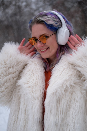 Cheerful woman in sunglasses walking outdoors, on the street on a winter day. A woman with bright multi-colored hair listens to music using wireless headphones.の写真素材