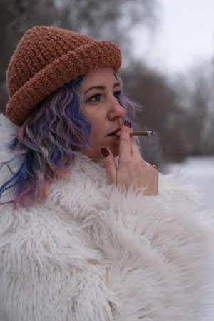 A woman with bright colored hair, wearing a white fur coat, smokes a cigarette outdoors in winter. Young woman in casual urban styleの写真素材