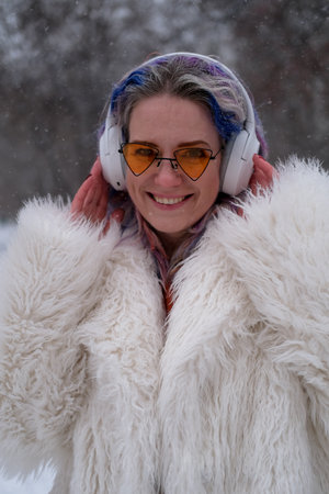 Cheerful woman in sunglasses walking outdoors, on the street on a winter day. A woman with bright multi-colored hair listens to music using wireless headphones.の写真素材
