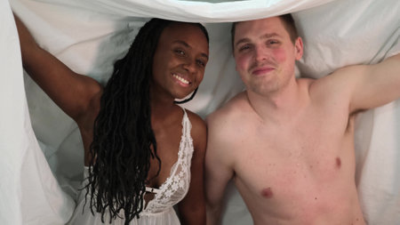 Sensual multiethnic couple kissing and hugging in bed at home. Romantic guy and girl having a good time together in the bedroom. A beautiful wife and a handsome husband look at each other, enjoy a tender moment. Happy loving couple.の写真素材