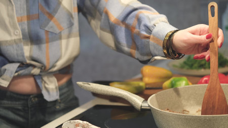 A woman is preparing dinner in the kitchen, stirring vegetables in a frying pan with a wooden spatula. View against the background of fresh bell pepper lying on a cutting boardの写真素材