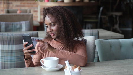 Cute African American woman drinking coffee or tea and using mobile phone in cafe