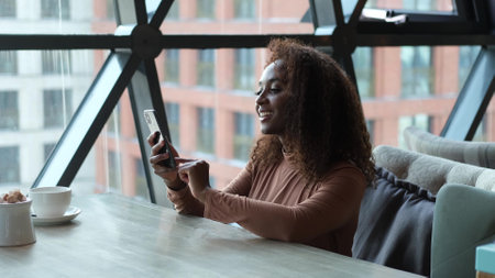 Cute African American woman drinking coffee or tea and using mobile phone in cafe