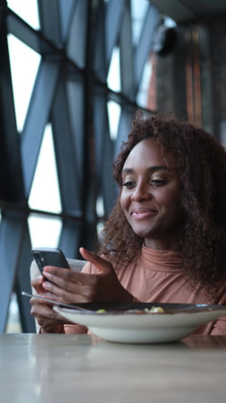 Cute African American woman drinking coffee or tea and using mobile phone in cafe