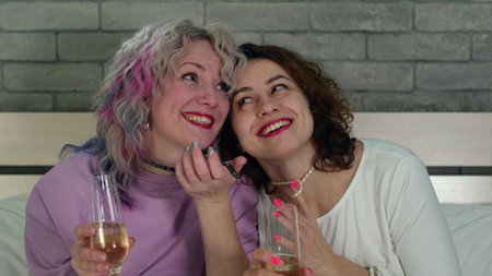 Two women with curly hair and bright makeup drink champagne in the bedroom at home and have fun