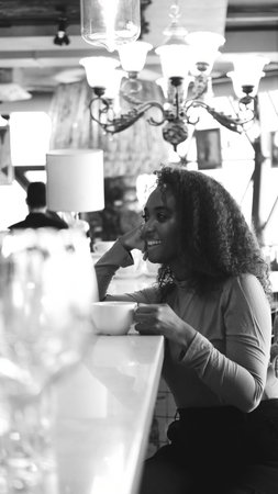 A very beautiful African American woman is sitting at a table in a cafe with a cup of coffee or tea.
