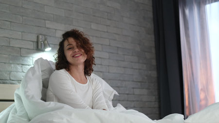 Happy sleepy young adult woman waking up, sitting in comfortable bed early in the morning. Positive beautiful girl stretches enjoying early good morning in cozy bedroom. Fresh lazy lady waking up at home after healthy sleepの写真素材