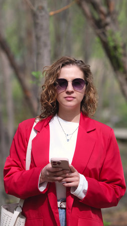 A charming curly young woman in a red jacket reads a message on her mobile phone against the...