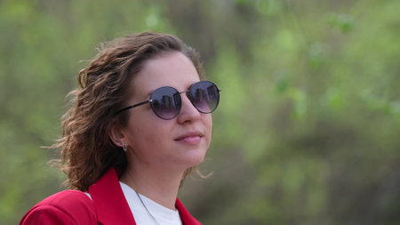A young woman with curly hair in a red jacket walks through a spring park, resting from a working...