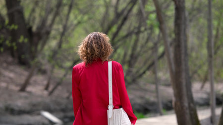 A young woman with curly hair in a red jacket walks through a spring park, relaxing after a working...