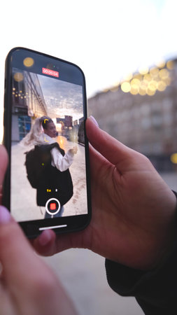 A girl with long white hair poses while filming a video on a smartphone against the backdrop of the...