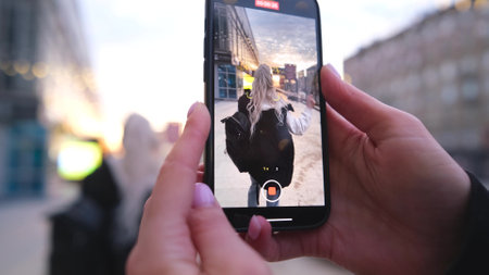 A girl with long white hair poses while filming a video on a smartphone against the backdrop of the setting sun on a city street.の写真素材