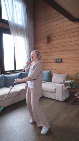 Happy young woman singing while washing floor at living room, using mop as microphone, having fun during house chores. Millennial lady enjoying domestic duties, gamifying cleaning routineの写真素材