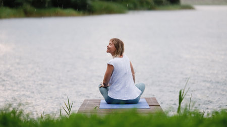 Young Caucasian woman sitting in lotus position on bridge by lake during rain. Woman enjoying fresh air. Outdoor meditationの写真素材