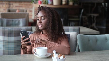 Cute African American woman drinking coffee or tea and using mobile phone in cafe