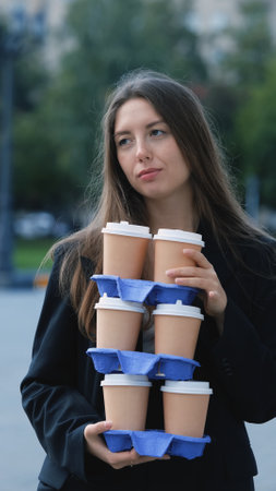 Pretty woman with long hair in office black jacket walking to work carrying many cups of takeaway coffee in her handsの写真素材