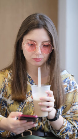 Pretty brunette with phone in hands relaxing in cafe. Woman drinking coffee cocktail