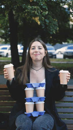 Beautiful woman with long hair in office clothes sits on a bench in a city park, holding many cups...
