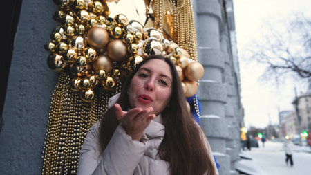 Portrait of a beautiful young woman in winter clothes. The woman smiles happily against the background of a snowy street traditionally decorated for Christmas holidaysの写真素材