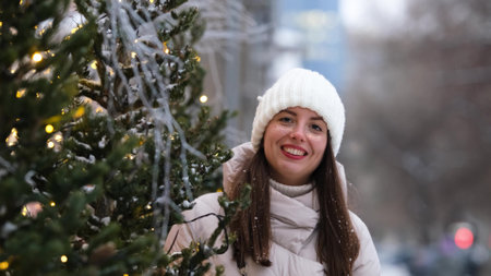Portrait of a beautiful young woman in winter clothes. The woman smiles happily against the background of a snowy street traditionally decorated for Christmas holidaysの写真素材