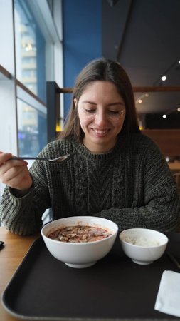 Pretty brunette enjoying delicious Tom Yum Kun soup with shrimps in Thai restaurant winter in Siberia. A woman in a warm cozy sweater is having lunch near the window. Snow outsideの写真素材
