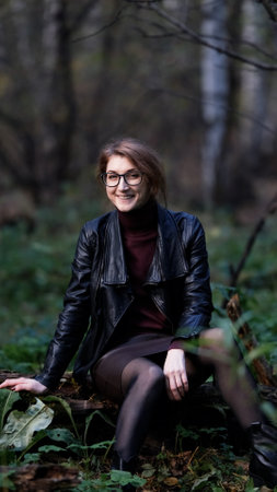 Smiling city woman relaxing in the forest. Thoughtful woman in glasses and office clothes sitting...
