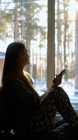 Beautiful young woman sitting on the floor near a large window with a smartphone in her hands while...