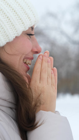 Woman in warm white knitted hat rubbing frozen hands to warm them up. Active winter recreation. Wide, beautiful smile. Close-upの写真素材