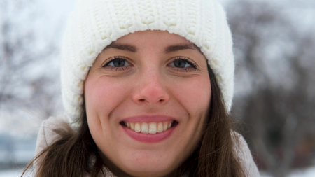 Portrait of woman in warm white knitted hat outdoors in winter. Young woman smiling and enjoying...