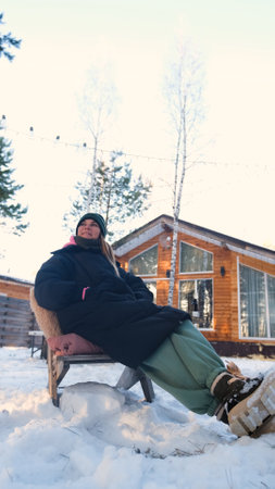 A woman in warm winter clothes sits in an outdoor chair in winter surrounded by snowdrifts near her...