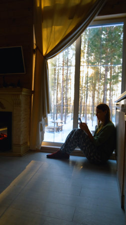 Young woman sitting on the floor near a large window with a smartphone in her hands while relaxing...