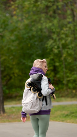 Beautiful blonde woman walking through city park holding roller skates enjoying beautiful day