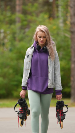 Beautiful blonde woman walking through city park holding roller skates enjoying beautiful day