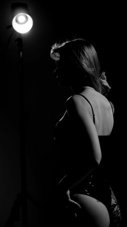 Blonde with a beautiful figure in a black patent leather bodysuit posing in a studio. Studio lighting, low exposure, monochrome imageの写真素材