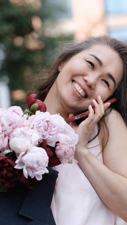 Beautiful woman with bouquet of peonies talking on phone and looking happy outdoors. Urban lifestyle concept.の写真素材