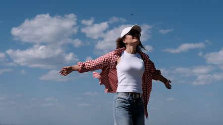 Young joyful woman in white baseball cap and red checkered shirt spinning on the seashore enjoying summer holidays. Vacation, weekend, holidaysの写真素材