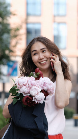 Beautiful woman with bouquet of peonies talking on phone and looking happy outdoors. Urban lifestyle concept.の写真素材