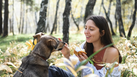 Cute young woman with her dog enjoying the beauty of nature and bonding with her pets. Woman eating bun and feeding her dog during outdoor picnic in autumn forest.の写真素材